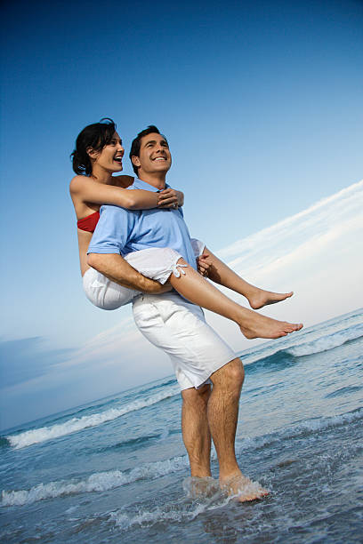 Man carrying woman piggyback at beach. Smiling caucasian mid-adult male carrying female on his back on beach. bald head island stock pictures, royalty-free photos & images