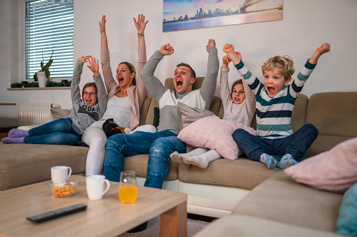 Front shot of excited white parents and kids seated at living room sofa. They have their arms up acting cheerful.