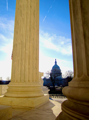 Washington, D.C., USA - February 17, 2017: The dome of the U.S. Capitol can be seen in the distance between columns of the U.S. Supreme Court on a sunny afternoon.
