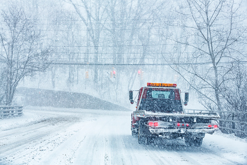A large flatbed emergency services vehicle tow truck is driving slowly and deliberately toward a slippery, sloppy rural red light traffic signal road intersection during a raging winter blizzard snow storm.
