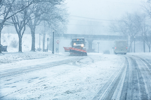 Winter snowplow dump truck clearing the road and spreading road salt is approaching on a slushy, slippery village street during a blizzard snow storm.