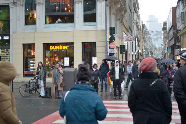 Brussels, Belgium - December 22, 2022: shoppers waiting for a green light to cross the street in the rain at the corner of a Dunkin patisserie