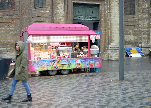 Brussels, Belgium - December 22, 2022: illuminated popcorn and candy pink market stall trailer in front of the church on a square. Shop owner looking suspicious to the camera