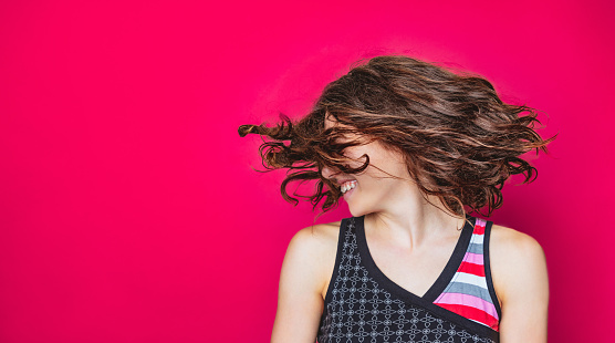 Portrait of young woman in sleeveless top smiling and tossing her hair turning head left on vivid magenta background