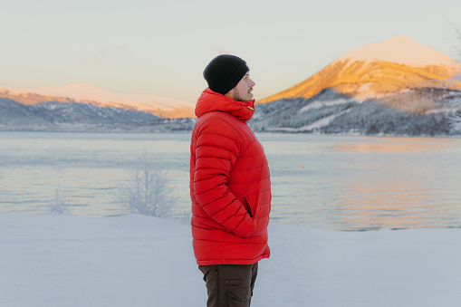 Happy male in red jacket walking at the frozen lakeshore admiring the sunset mountains in Scandinavia
