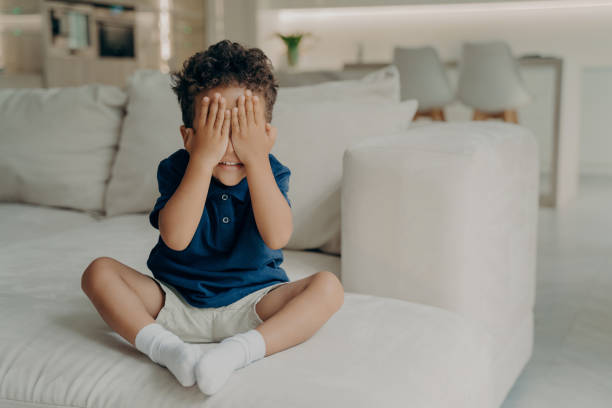 Small excited boy in tshirts and shorts playing game of hide and seek, covering face with his hands, eager to start searching, waiting and counting while sitting on white couch in light modern room