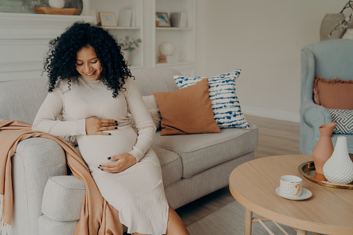 Smiling lovely pregnant mixed race woman in white dress with curly hair sitting on couch in front of round table and thinking about motherhood, happy to become mother. Pregnancy concept