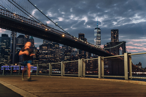 A man is running in New York City - Skyline at night with Brooklyn Bridge and Lower Manhattan in the background. Image shot from Brooklyn.