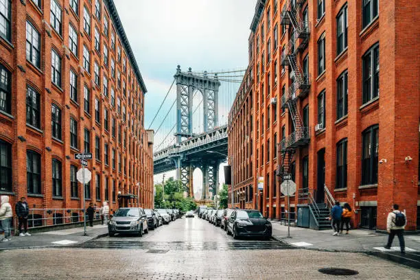 Dumbo district view in Brooklyn with Manhattan Bridge and New York City in the background. Raining day in NYC. There is a rainbow in the cloudy sky.