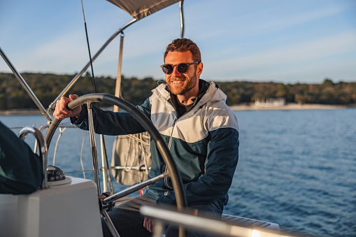 Waist up shot with blurred background of a good looking sailor smiling and wearing sunglasses while holding the boat helm and looking and enjoying at the sunset on a summer day in the Adriatic sea.