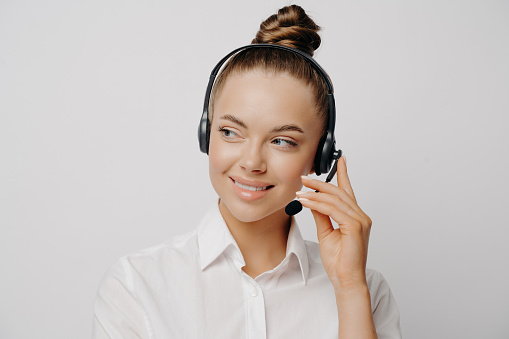 Close up portrait of female bank worker in white shirt and hair in bun, speaking on black headset, telling customer information and looking aside with smile isolated over grey background