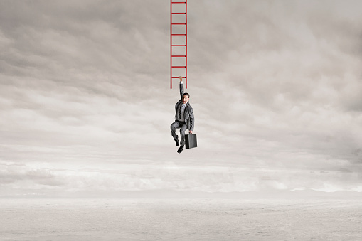 A man holding a briefcase hangs by one arm from the bottom rung of a red ladder. The man looks down in fear as he wonders how he is going to safely reach the ground.