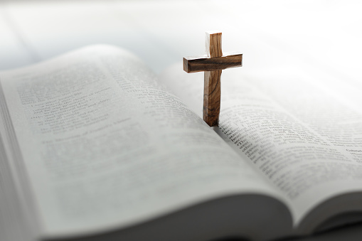 An open holy bible on a white wooden surface with a wooden cross standing upright on its open pages. Focus on the cross with the pages of the book largely defocused.