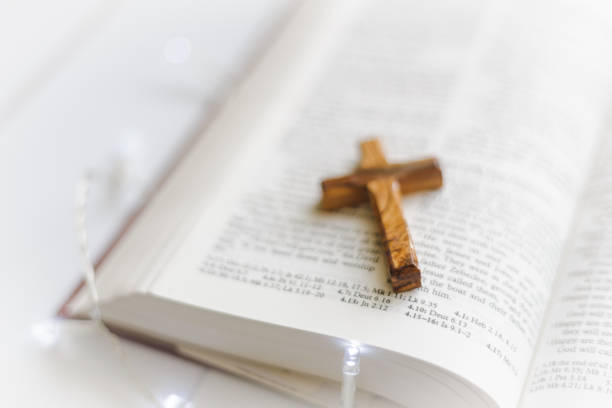 An open holy bible on a white wooden surface with a wooden cross lying on its open pages. Focus on the cross with the pages of the book largely defocused.