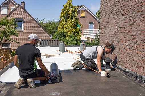 Urk, The Netherlands - June 23, 2022: Workers at flat roof preparing rolls of new white roofing felt or bitumen
