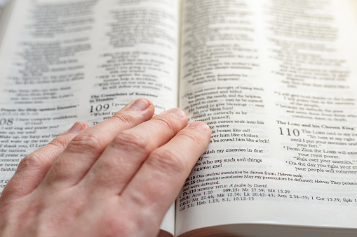 An open holy bible on a white wooden surface with a human hand resting on it. Focus on the cross with the pages of the book largely defocused.