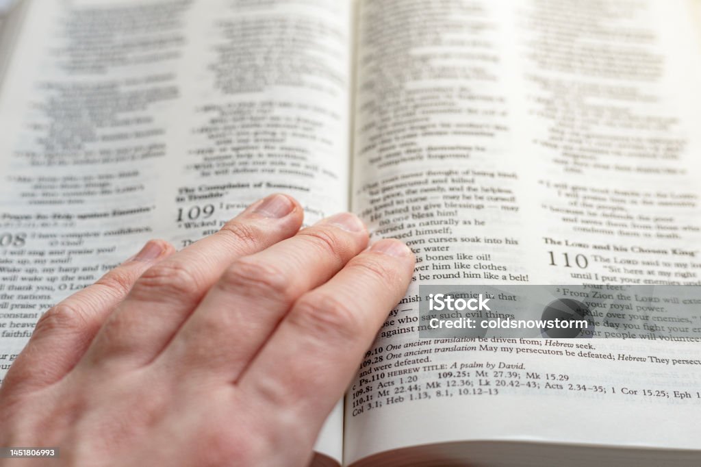Hand resting on pages of holy bible An open holy bible on a white wooden surface with a human hand resting on it. Focus on the cross with the pages of the book largely defocused. Bible Stock Photo