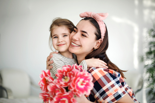 Smiling brunette mother hugging little daughter in cozy bedroom at weekend celebration Happy Mothers Day holding blooming bouquet of tulips