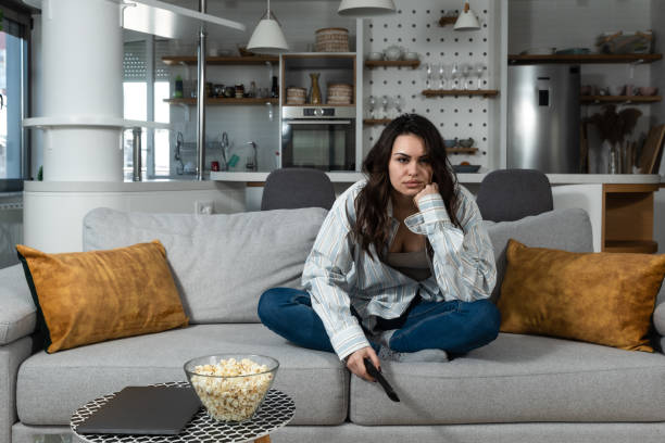 Young freelancer business woman bored by tv program sitting at home on day off dont know what to do. Casual female feel boredom at apartment during her sick days eating popcorns and watch television. stock photo