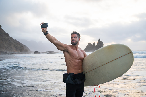Athletic young surfer takes a selfie, holding a surfboard and standing at the beach during sunset.