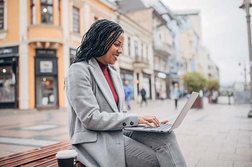 Modern black woman sitting on bench and using laptop on the street in city.