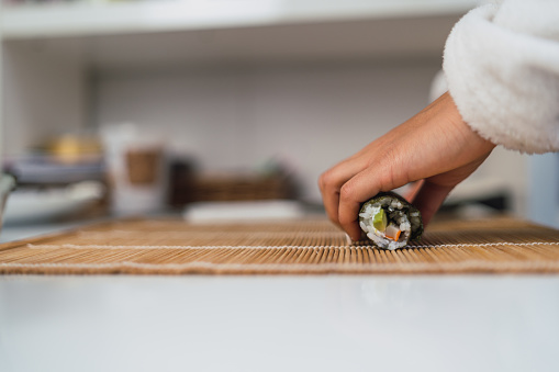 Close-up side image of an unrecognizable woman trying to roll with her hands a sheet of nori seaweed she is using to prepare homemade sushi at her kitchen table.