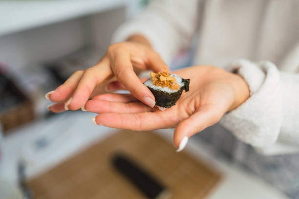 Unrecognizable woman's hands holding a slice of homemade sushi with crispy onion that she has just prepared in her kitchen.