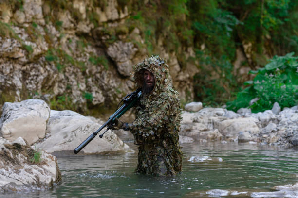 迷彩服を着た軍人またはエアガン奏者が川をこっそりと歩き回り、スナイパーライフルから横または標的を狙う。 - wet spy men suit ストックフォトと画像