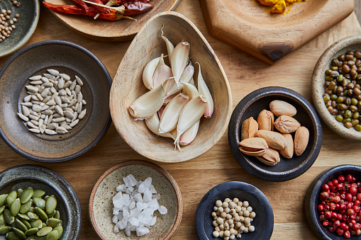 Variety of spices, herbs, nuts, seasoning, in a handmade wooden and ceramic bowls, on a kitchen cutting board, on an oak wooden table top, a top view macro photo shoot texture background, pumpkin seed, coriander seed, sunflower seed, pistachio, sea salt, garlic cloves, chili pepper and red pepper