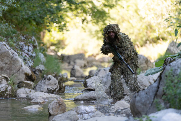 迷彩服を着た軍人またはエアガン奏者が川をこっそりと歩き回り、スナイパーライフルから横または標的を狙う。 - wet spy men suit ストックフォトと画像