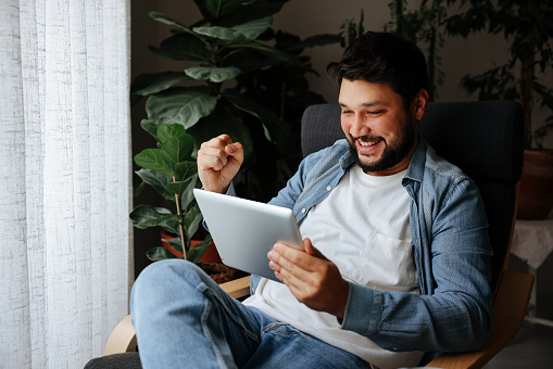 Young adult man holding coffee cup while looking away. Male is sitting on sofa at home. He is in casuals. Shot of a happy young man using his tablet while relaxing on the couch at home
