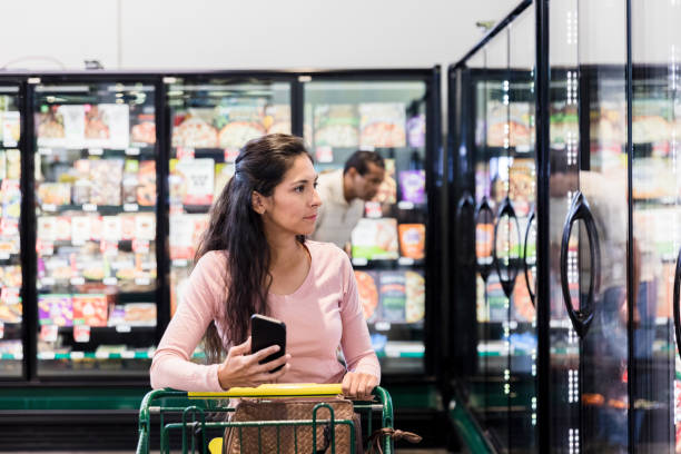 une femme utilise une application lorsqu’elle fait ses courses à l’épicerie - decisions women thinking latin american and hispanic ethnicity photos et images de collection