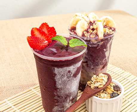 Two disposable cups with açaí on a jute surface, with strawberries, banana and granola. Brazilian popular food.