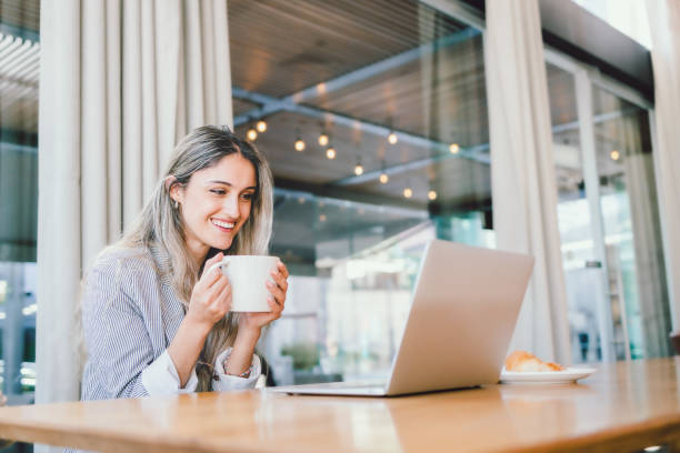 istock Young cheerful businesswoman using her laptop , drinking coffee from a mug in modern open workplace cafe. 1451850986