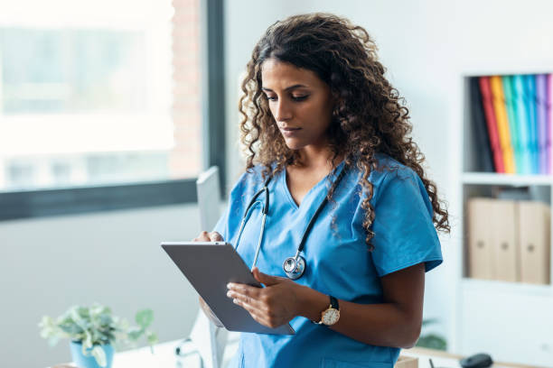 Female nurse using her digital tablet while standing in the consultation. Shot of female nurse using her digital tablet while standing in the consultation. nurse stock pictures, royalty-free photos & images