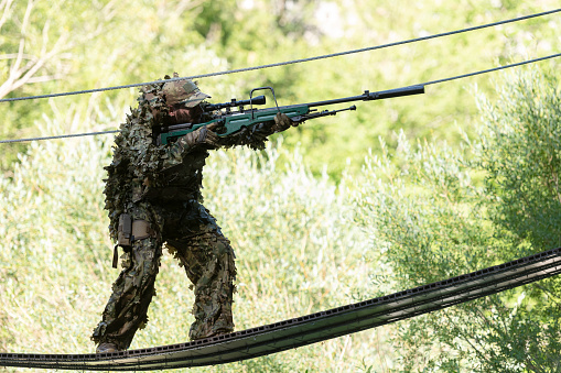 A military man or airsoft player in a camouflage suit sneaking the rope bridge and aims from a sniper rifle to the side or to target.