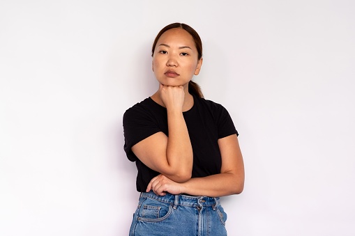 Portrait of serious young woman posing with hand on chin over white background. Asian lady wearing black T-shirt and jeans looking at camera. Confidence and boredom concept