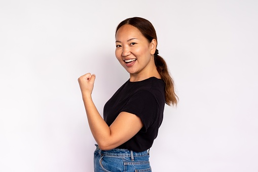 Portrait of self assured young woman making winning gesture over white background. Asian lady wearing black T-shirt and jeans showing bicep. Self esteem and strength concept