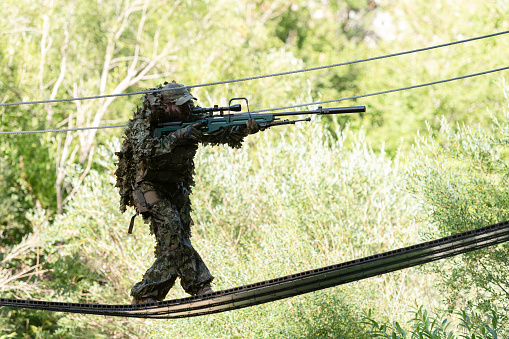 A military man or airsoft player in a camouflage suit sneaking the rope bridge and aims from a sniper rifle to the side or to target.