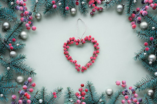 Christmas heart wreath made of berries. Frame with fir twigs and frosted red berries. Top view, flat lay, festive Xmas background on light mint green paper background.