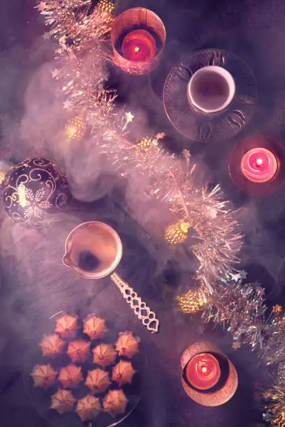Flat lay with copper cezve coffee pot, traditional bronze metal coffee cup, marzipan sweets, candles and light garland. Dark red flat lay on dark background with smoke and steam.