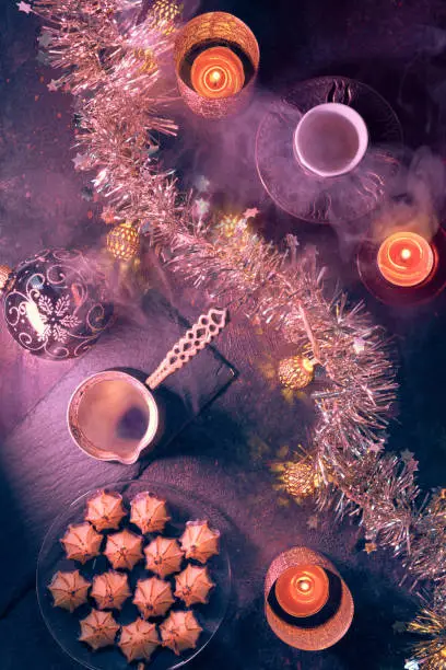 Flat lay with copper cezve coffee pot, traditional bronze metal coffee cup, marzipan sweets, candles and light garland. Dark red flat lay on dark background with smoke and steam.