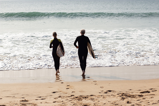 Back view of sporty senior couple holding surfboards. Grey-haired man and woman carrying boards going barefoot into beautiful sea for surfing. Sport activity, healthy lifestyle of aged people concept