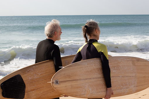 Back view of aged couple carrying big surfboards. Grey-haired man and woman in wetsuits walking along sea shore before starting surfing. Sport activity and healthy lifestyle of aged people concept