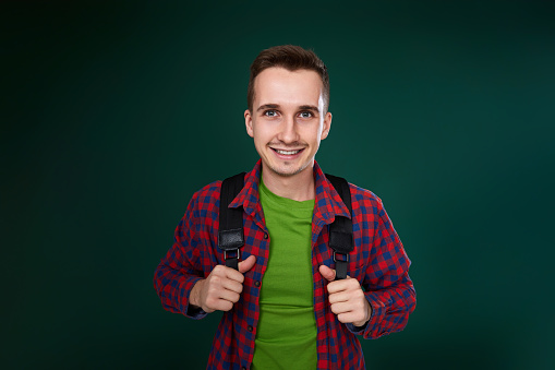 cheerful handsome man in red plaid shirt with backpack looking at camera on green background