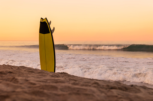 This is a photo of a yellow surfboard stuck in the sand on the beach by the ocean waves at the golden hour of sunset