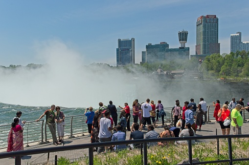 Niagara Falls, New York USA: May 28, 2018. Tourists observe the awe inspiring Niagara Falls at one of the many viewing platforms at the edge of the falls. The city of Niagara Falls Ontario Canada is seen on the other side of the river.