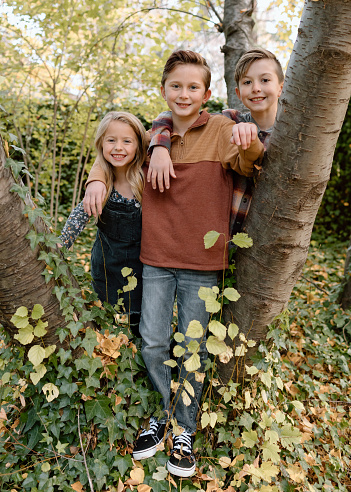A cute little girl and her two older brothers pose together and smile directly at the camera while playing together outside in nature on a crisp Autumn afternoon.