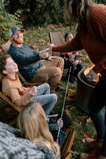 A family with elementary age children gathers around a fire while camping in the Autumn and roasts marshmallows.