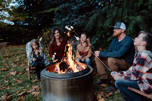 A man and woman with three elementary age children gather around a fire pit in the backyard and roast marshmallows on a cool Autumn evening. Selective focus on the fire and marshmallows.